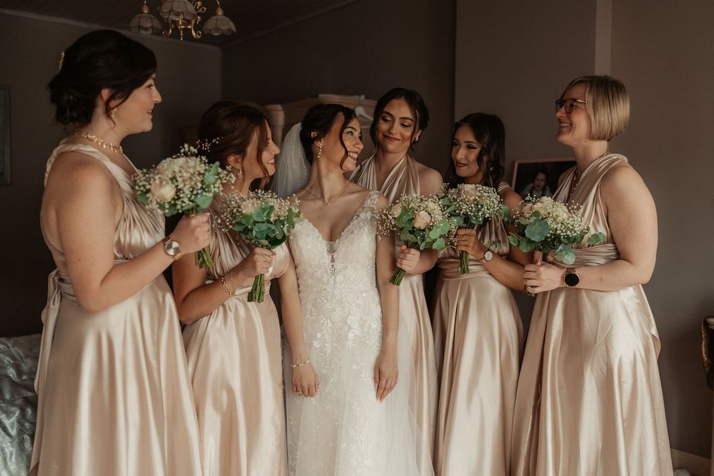 Bridesmaids in champagne dresses and bride in white lace gown pose with green bouquets in a soft-lit room.