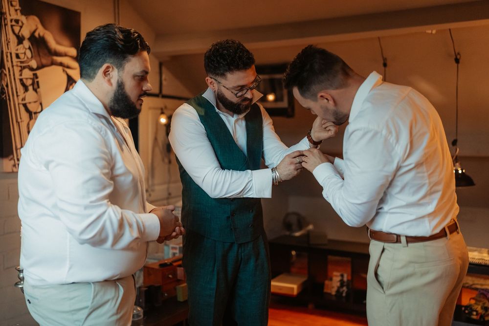 Three people in formal attire assist each other with getting ready for a special event in a warm-lit room.