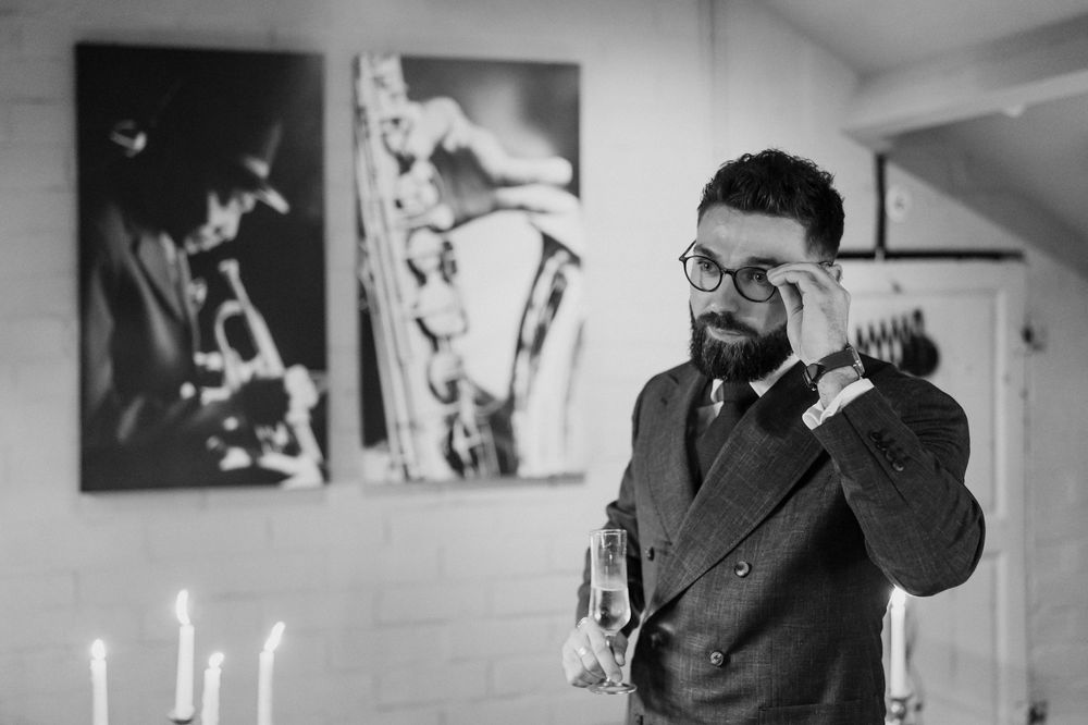 Black and white photo showing someone in a suit talking on phone with jazz-themed artwork on wall and candles in foreground.