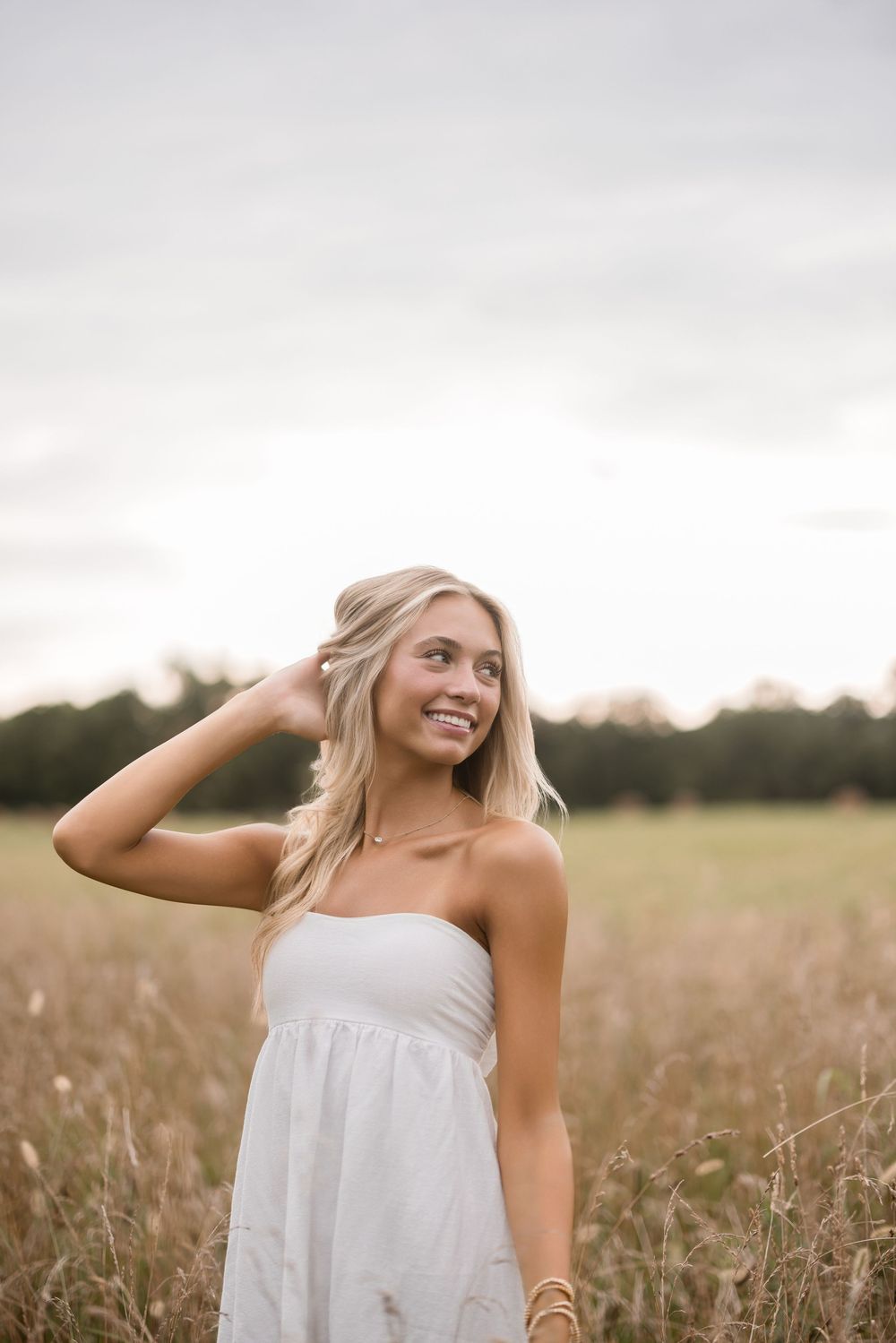 Person in white strapless dress poses in meadow during golden hour with windswept blonde hair.