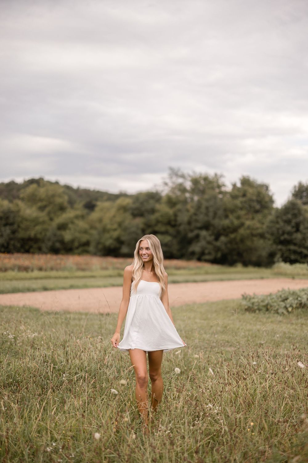 Person wearing white sundress stands gracefully in rustic field with wooded backdrop.