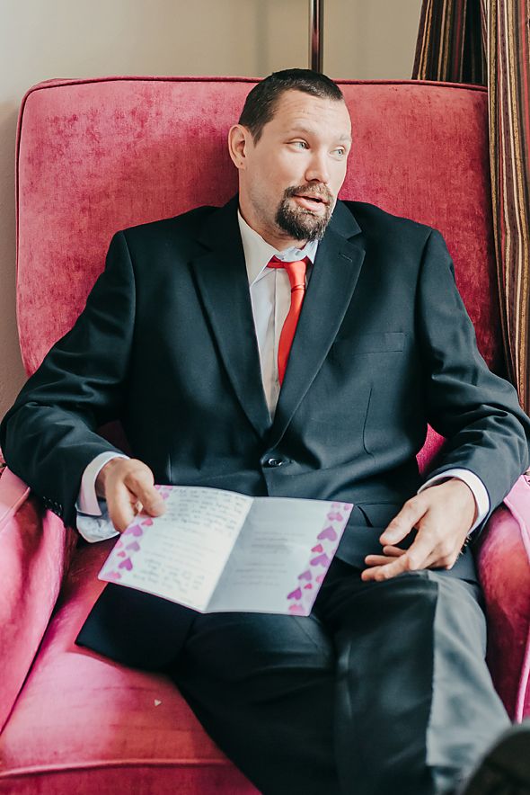 A person in a black suit and red tie sits in a pink armchair reading a document or notebook.