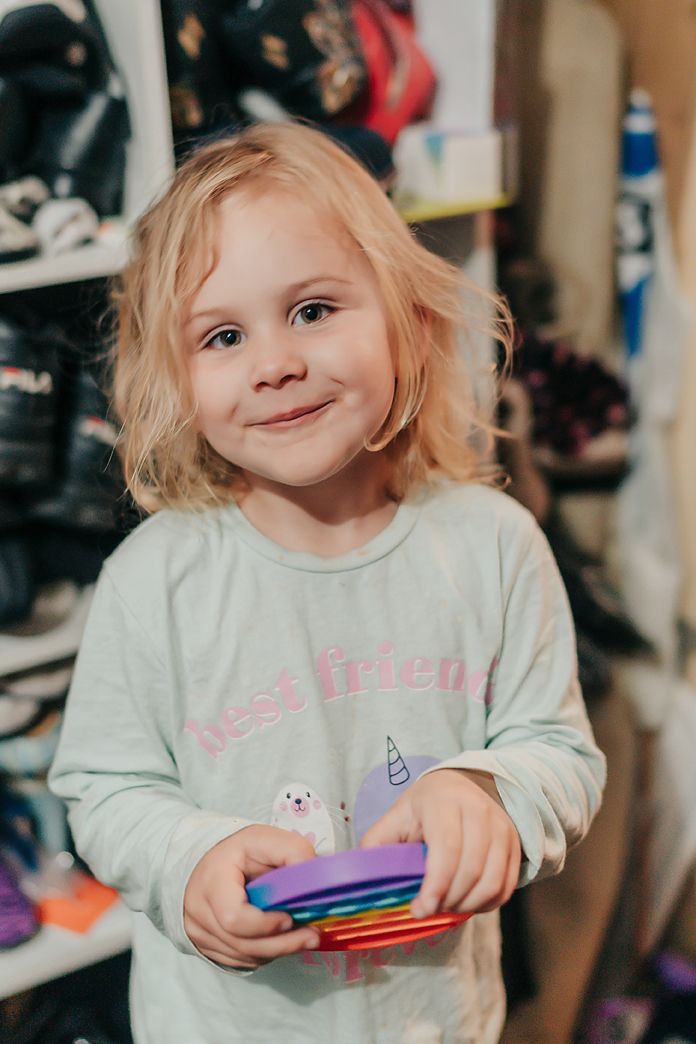 A smiling child with blonde wavy hair wearing a light mint sweatshirt holds colorful pens in a cluttered room.