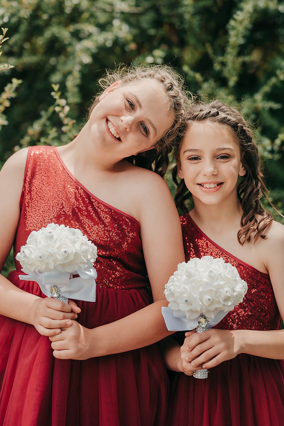 Bridesmaids in red sequin dresses hold white floral bouquets against a green leafy background during a wedding photo.