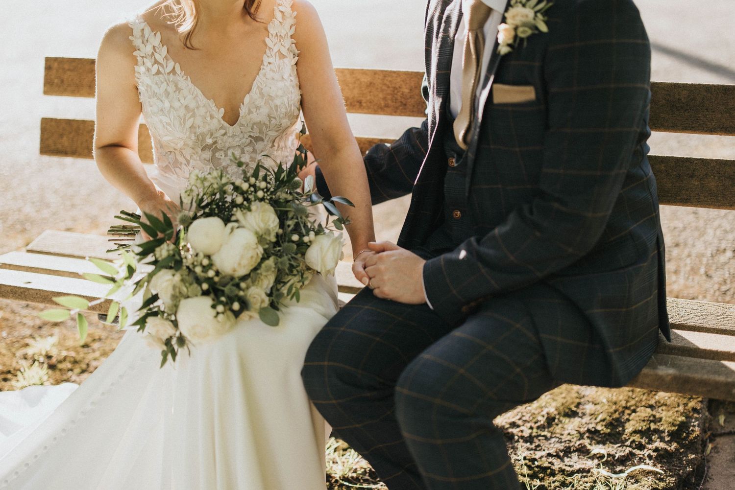 A couple sits on wooden bench during a wedding ceremony as bride holds elegant white floral bouquet.