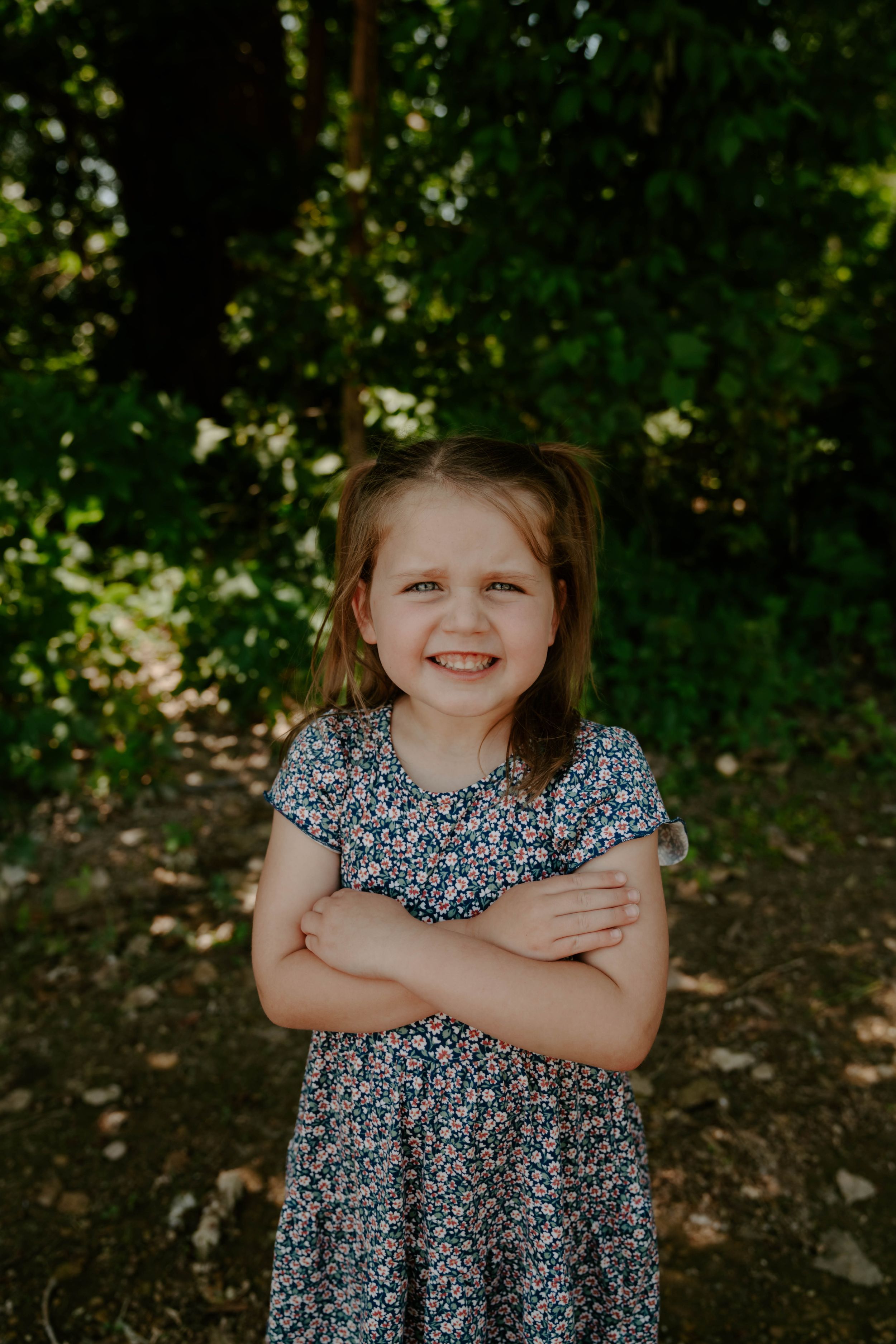 A child in a blue floral dress stands confidently with crossed arms in a forest setting.