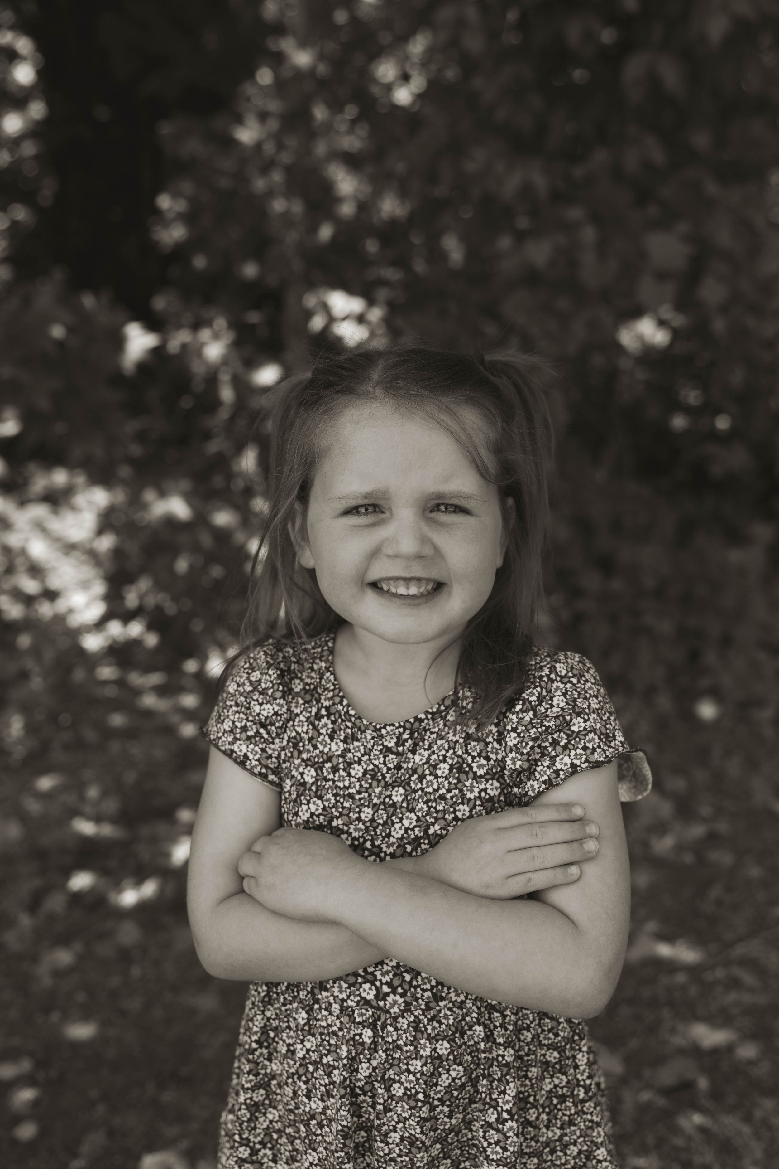 A candid black and white portrait of a child in a floral dress against a natural backdrop.