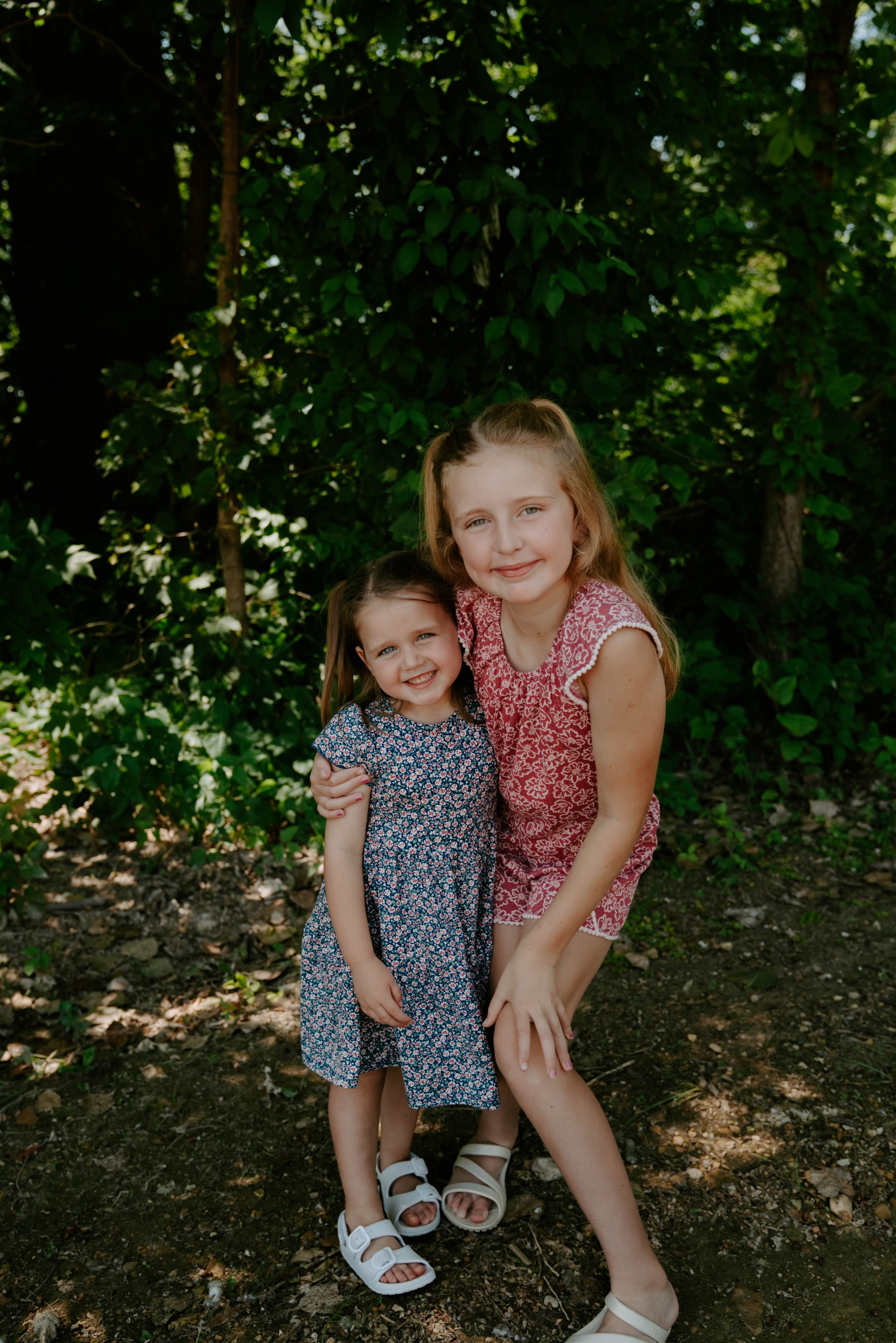 Two children in coordinating floral dresses sit together on the ground outdoors.