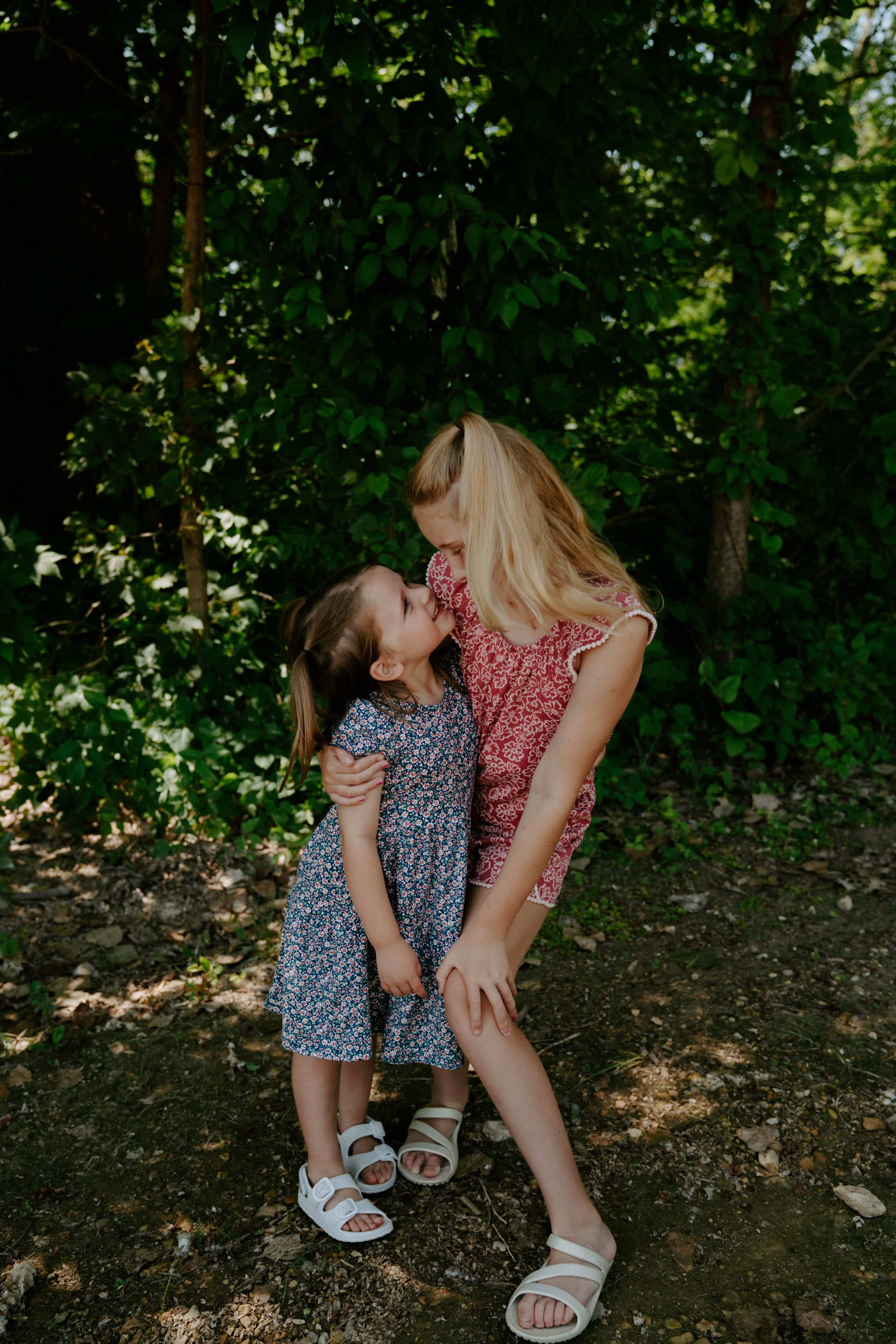 Two children in floral dresses share a playful moment while sitting on the ground.