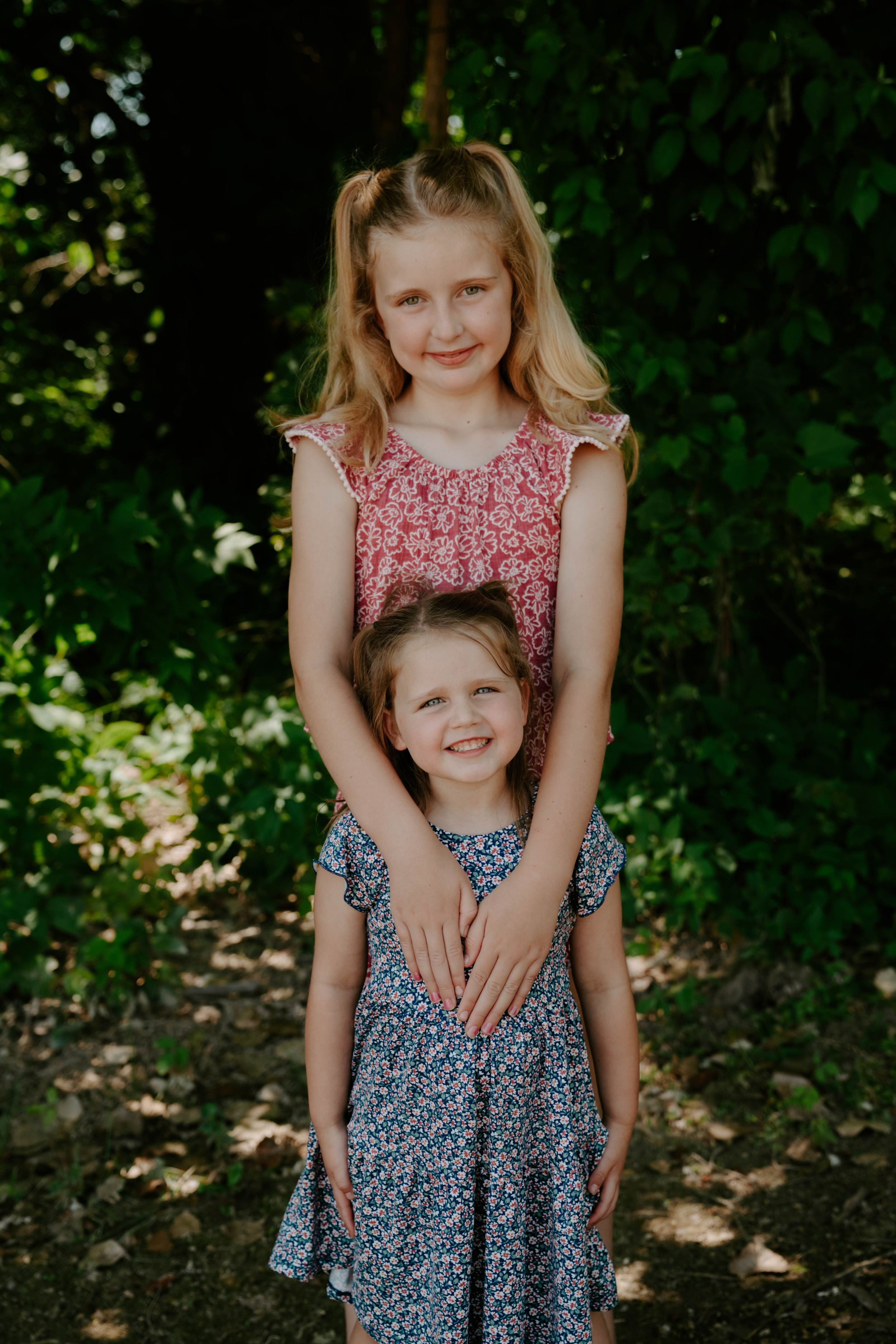 Two children in complementary floral dresses pose together in a wooded setting.