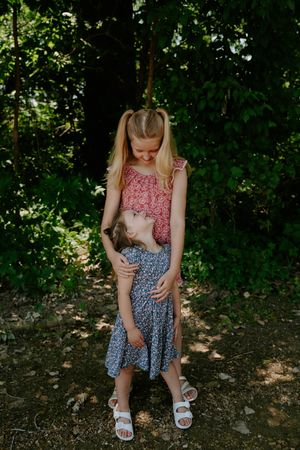 Two children in floral dresses share a tender moment in a forest setting.
