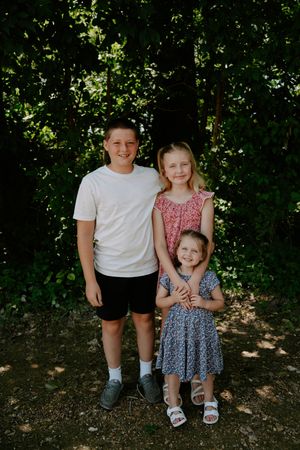 Three children pose together for a casual outdoor portrait in a wooded area.