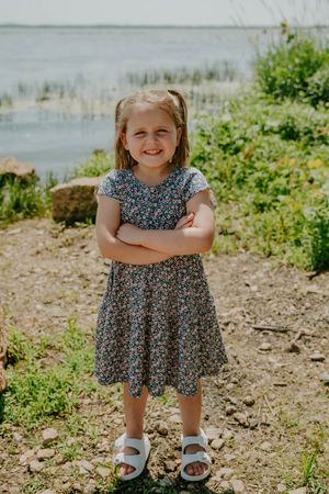 Young child in a floral dress poses confidently near a waterfront setting.