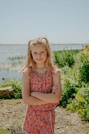 Child in red patterned dress stands cheerfully by the water's edge.