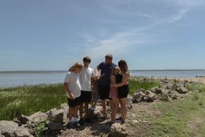A group explores a rocky shoreline with tall grass and water in the background.