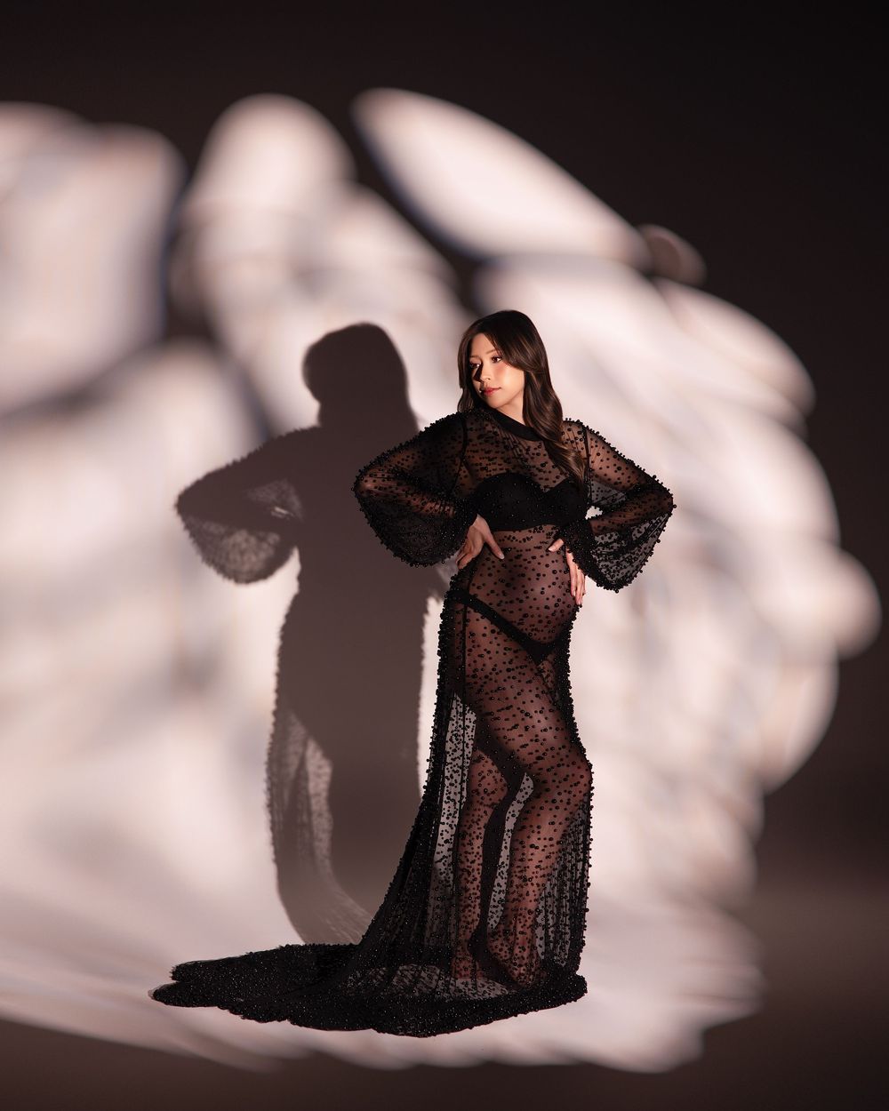 Pregnant woman in sheer black lace gown poses elegantly with dramatic light and shadow backdrop.