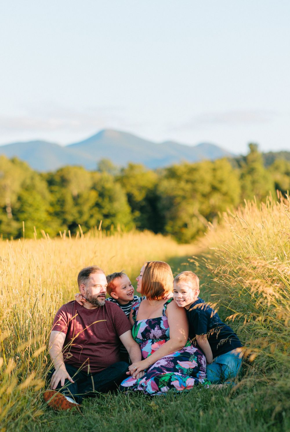 Family relaxes together in a golden wheat field at sunset with mountains visible in the distance.
