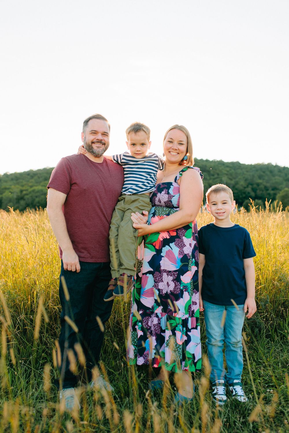 A happy family stands together in a golden wheat field at sunset with rolling hills in the background.