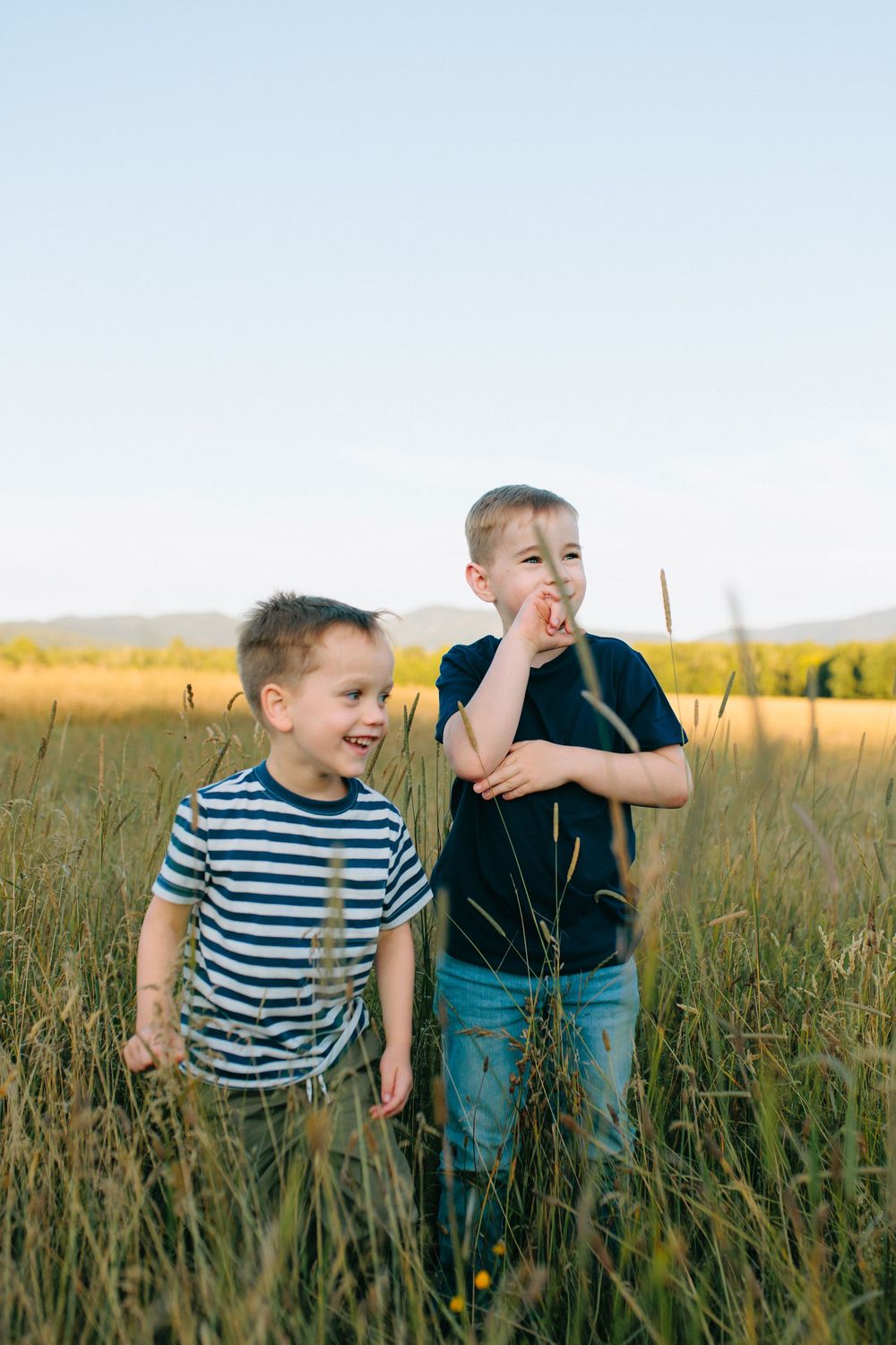 Two people stand together playfully in a grassy meadow during golden hour at sunset.