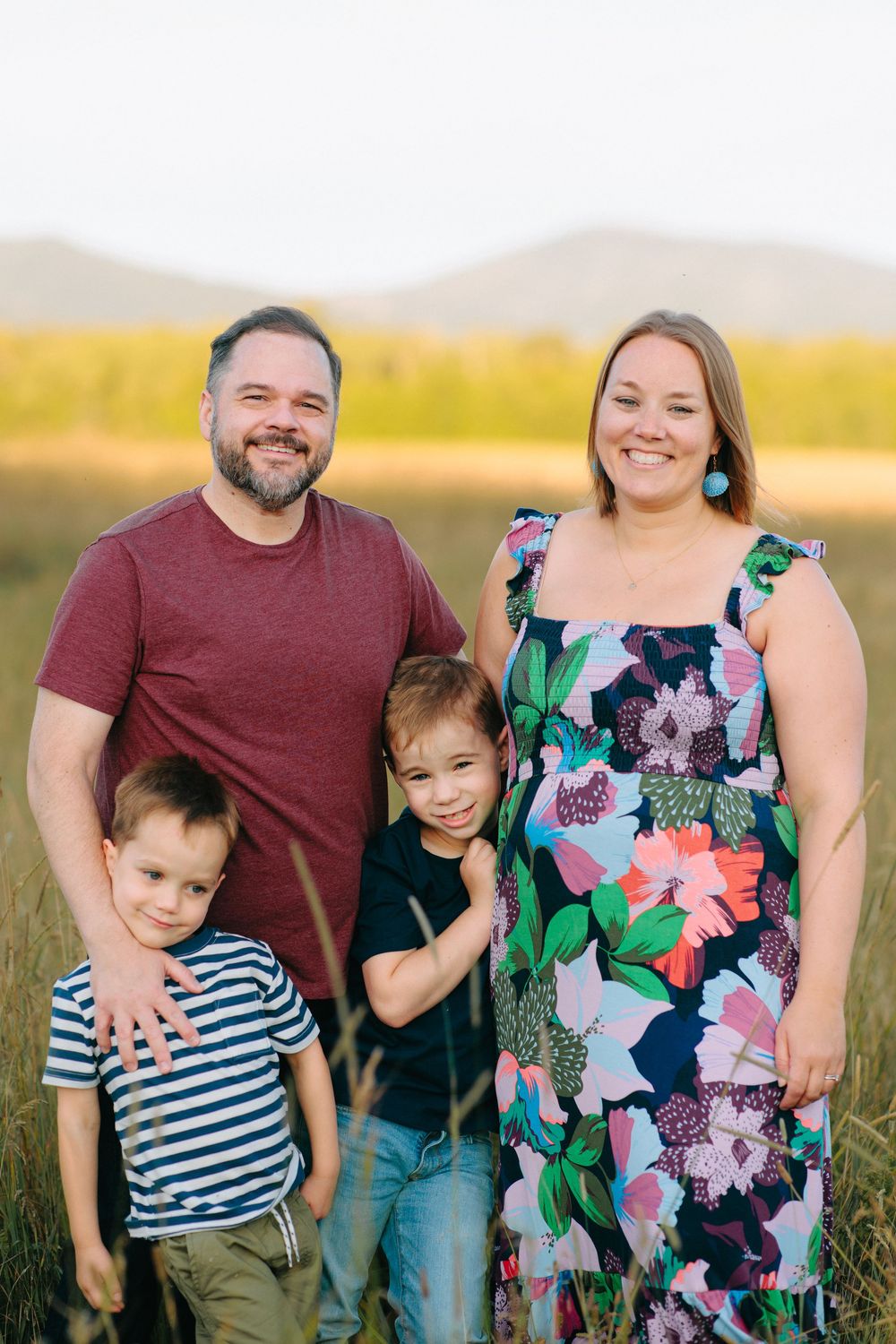 Family of four poses together in a grassy field during golden hour wearing coordinated casual outfits.