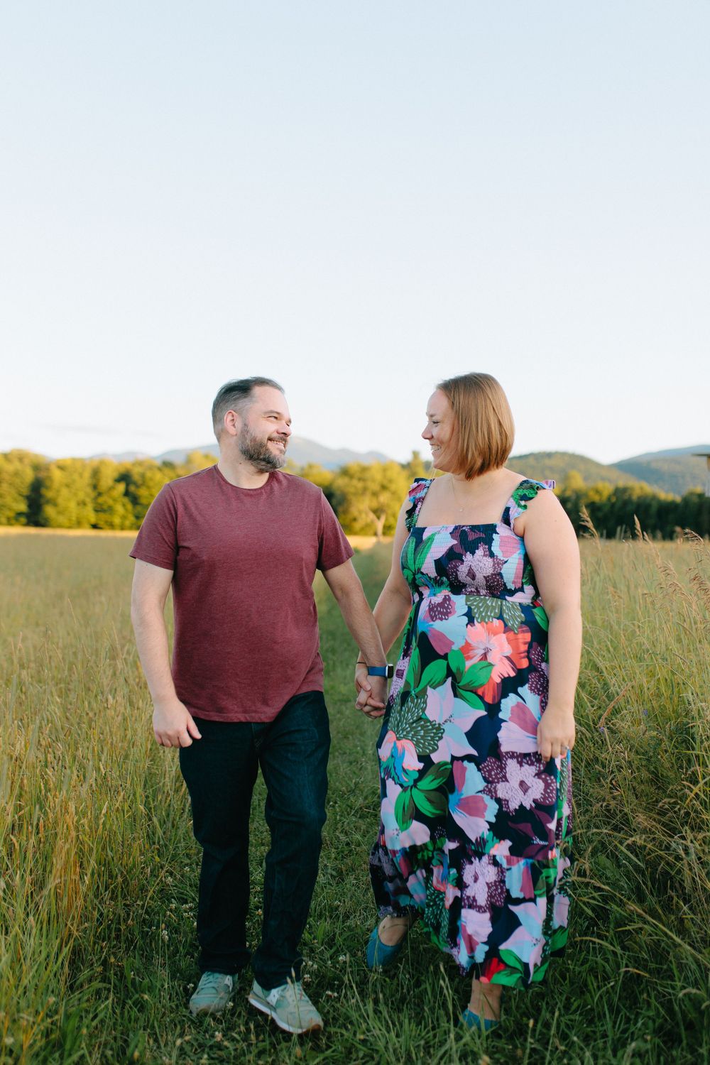 A couple holding hands while standing in a golden meadow at sunset with mountains in the background.