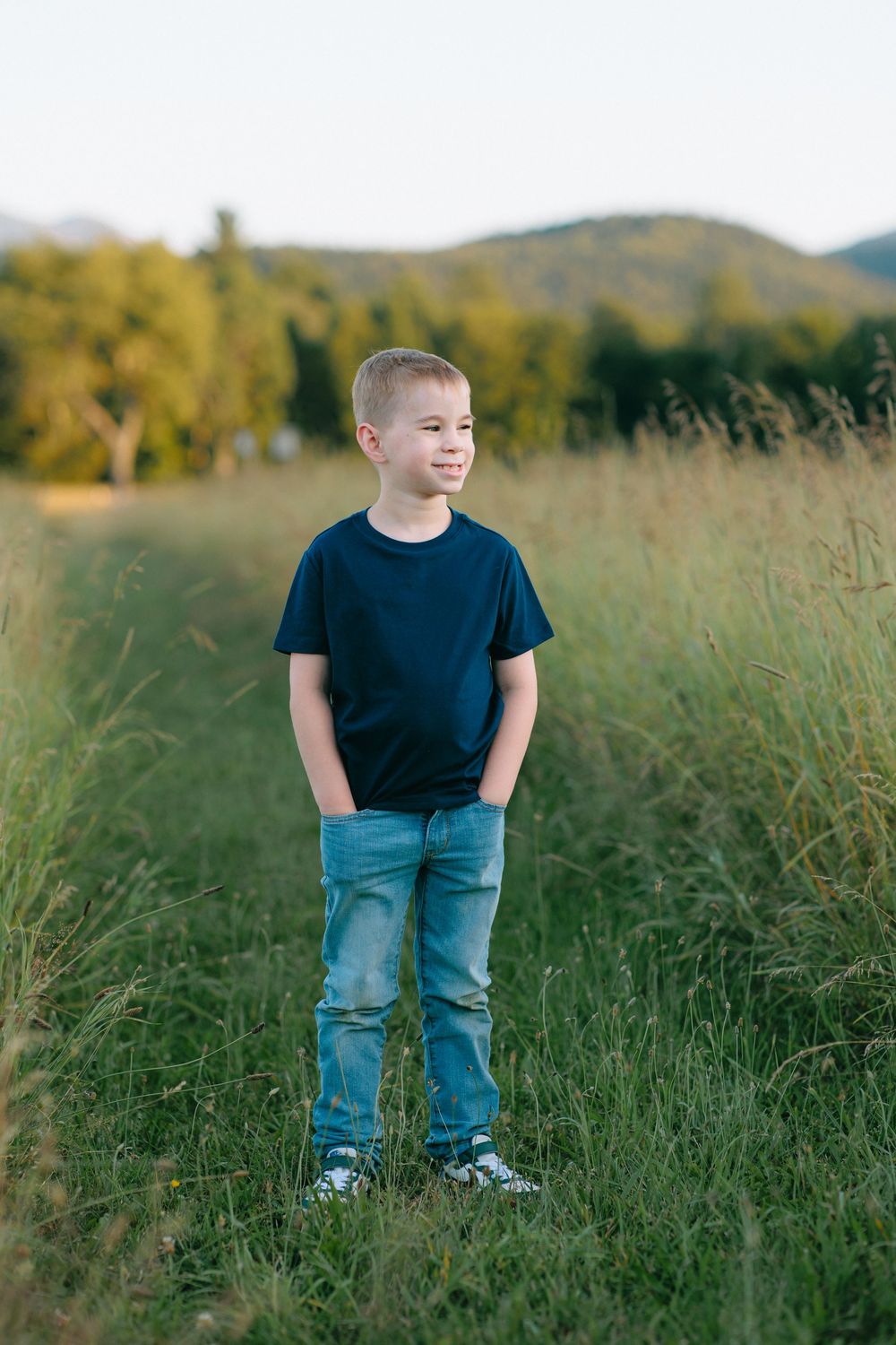 In a sunny field with mountains behind, a young child stands wearing a navy t-shirt and jeans among tall grass.