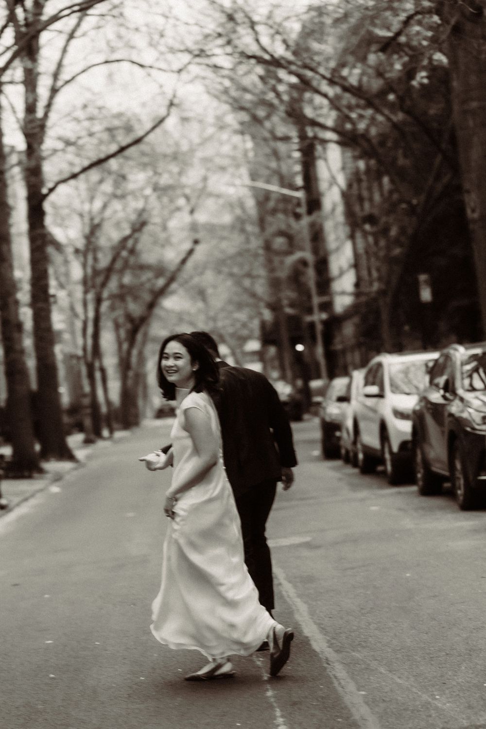A couple shares a joyful moment running across a tree-lined city street with parked cars in black and white.