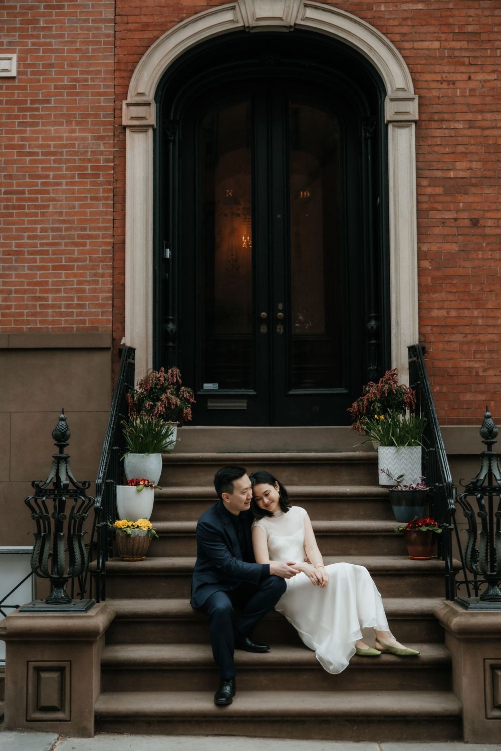 A couple sits on brick brownstone steps with a grand doorway decorated with potted flowers in New York City.