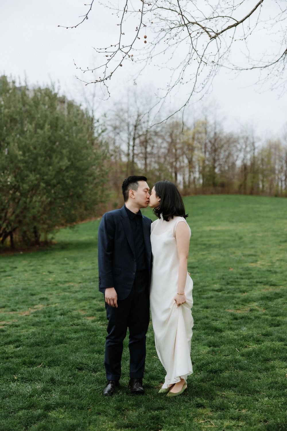 A couple in formal attire share an intimate moment on a green lawn with bare trees in the background.