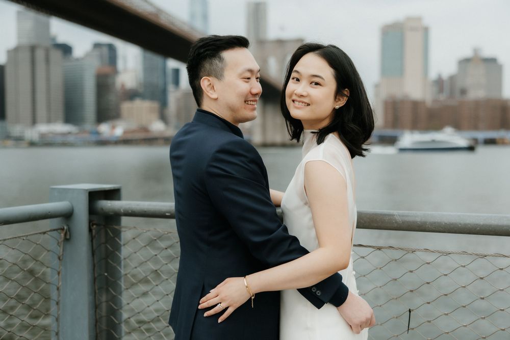 A couple shares an intimate moment by the waterfront with a city skyline backdrop.