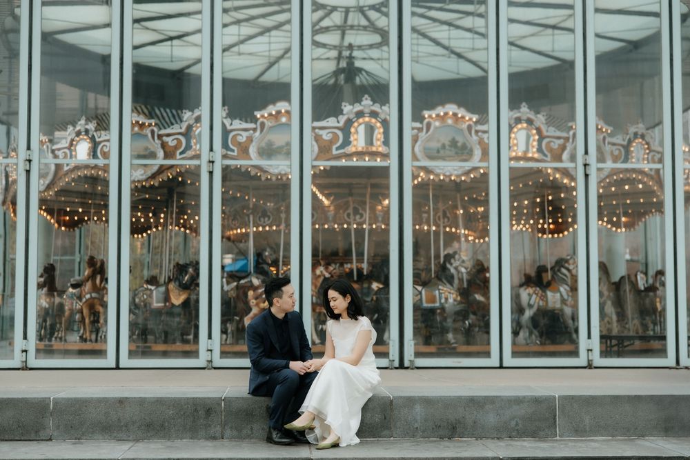 Wedding couple shares a romantic moment in front of floor-to-ceiling windows with ornate antique carousel horses behind.