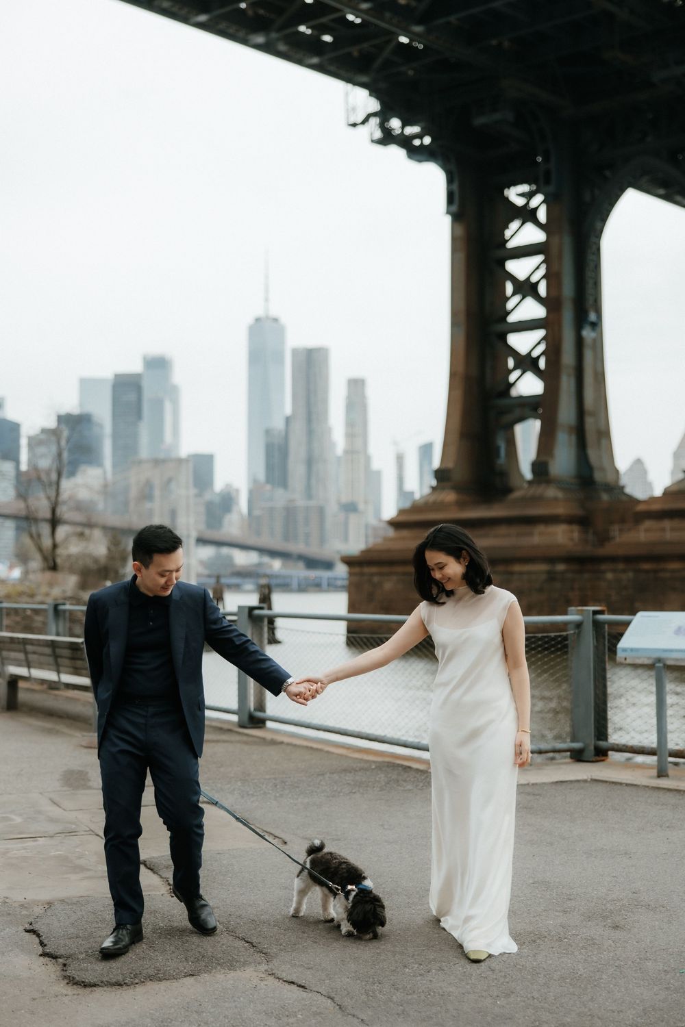 Under the bridge with New York skyline in the background, a couple shares a moment with their small dog in black and white.