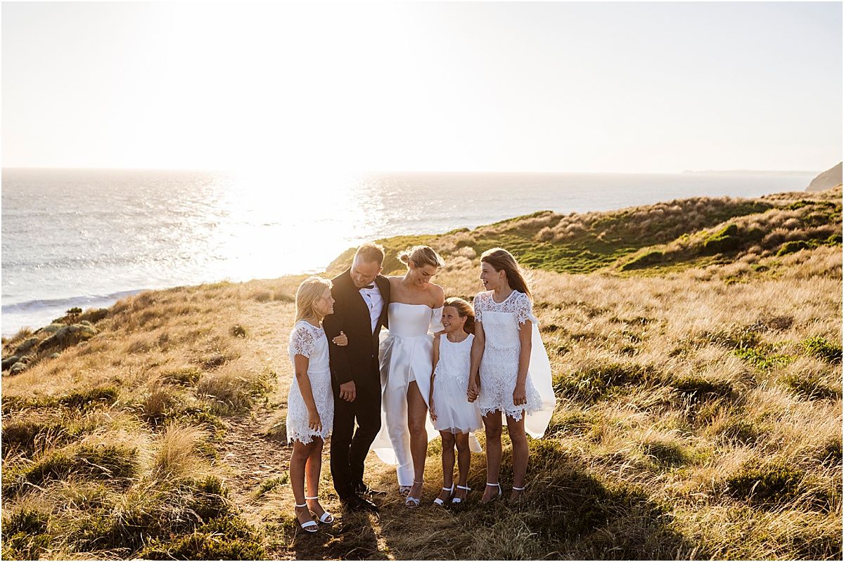 A wedding group stands together on a grassy ocean cliff at golden hour.