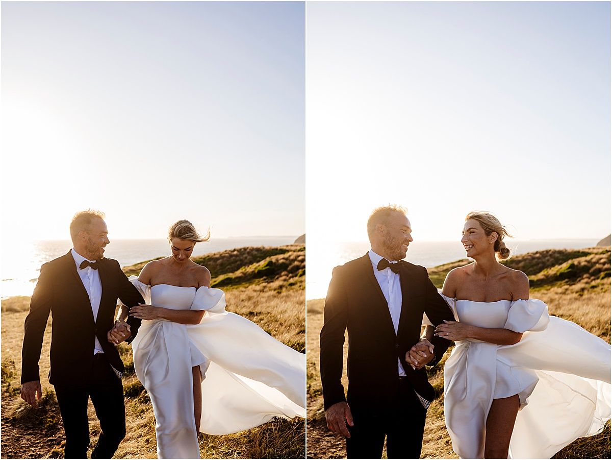 Couple in formal attire walking along coastal cliffs at sunset while bride's white dress flows in the wind.