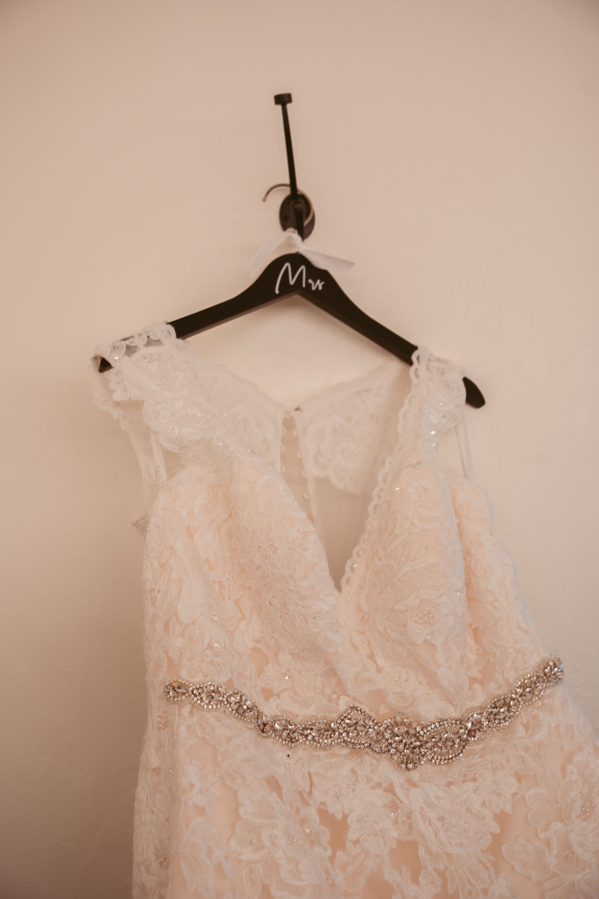 A delicate white lace wedding dress with crystal embellished belt hangs on a wooden Mrs hanger.