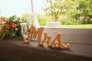 A wooden "Mrs" sign sits on a brown tablecloth with floral decorations and glassware at sunset.