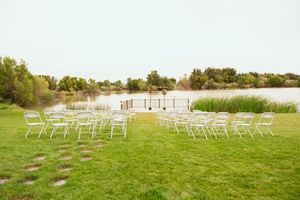White chairs arranged outdoors for a lakeside wedding ceremony on a green lawn next to a wooden deck.