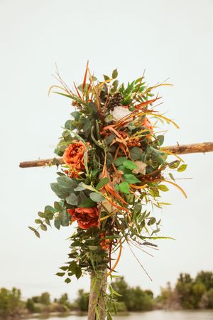 A rustic wooden cross decorated with orange and green floral arrangements featuring eucalyptus and roses.