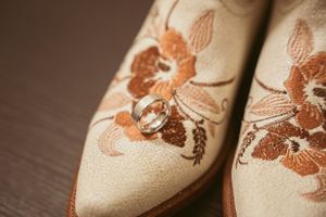 Close-up detail of beige leather boots with brown floral embroidery and a diamond ring placed on top.
