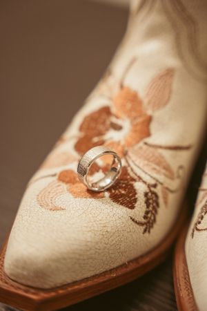 Wedding ring placed on embroidered cream-colored boot with floral copper pattern and leather sole.
