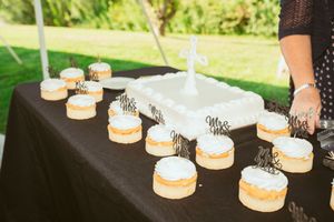 Simple white wedding cake and cupcakes arranged on black tablecloth at outdoor reception.