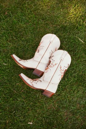 A pair of white leather cowboy boots with decorative stitching lies on green grass in various angles shown across four images.