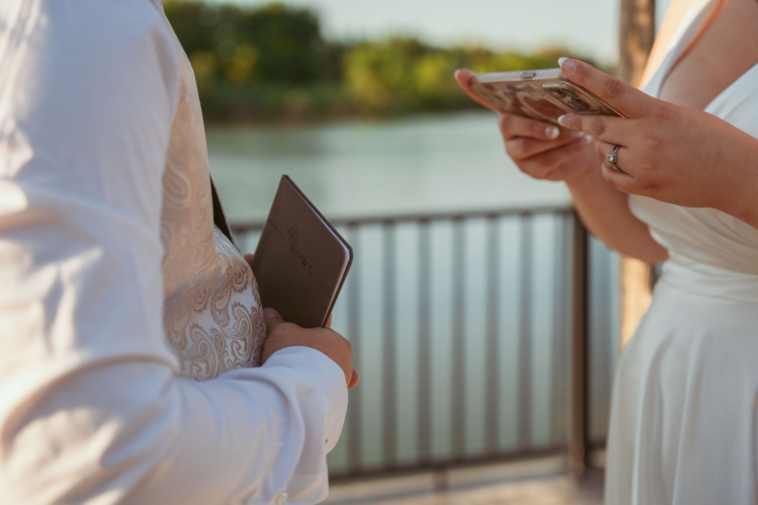 Couple exchanging vows or reading from small books during waterfront ceremony.