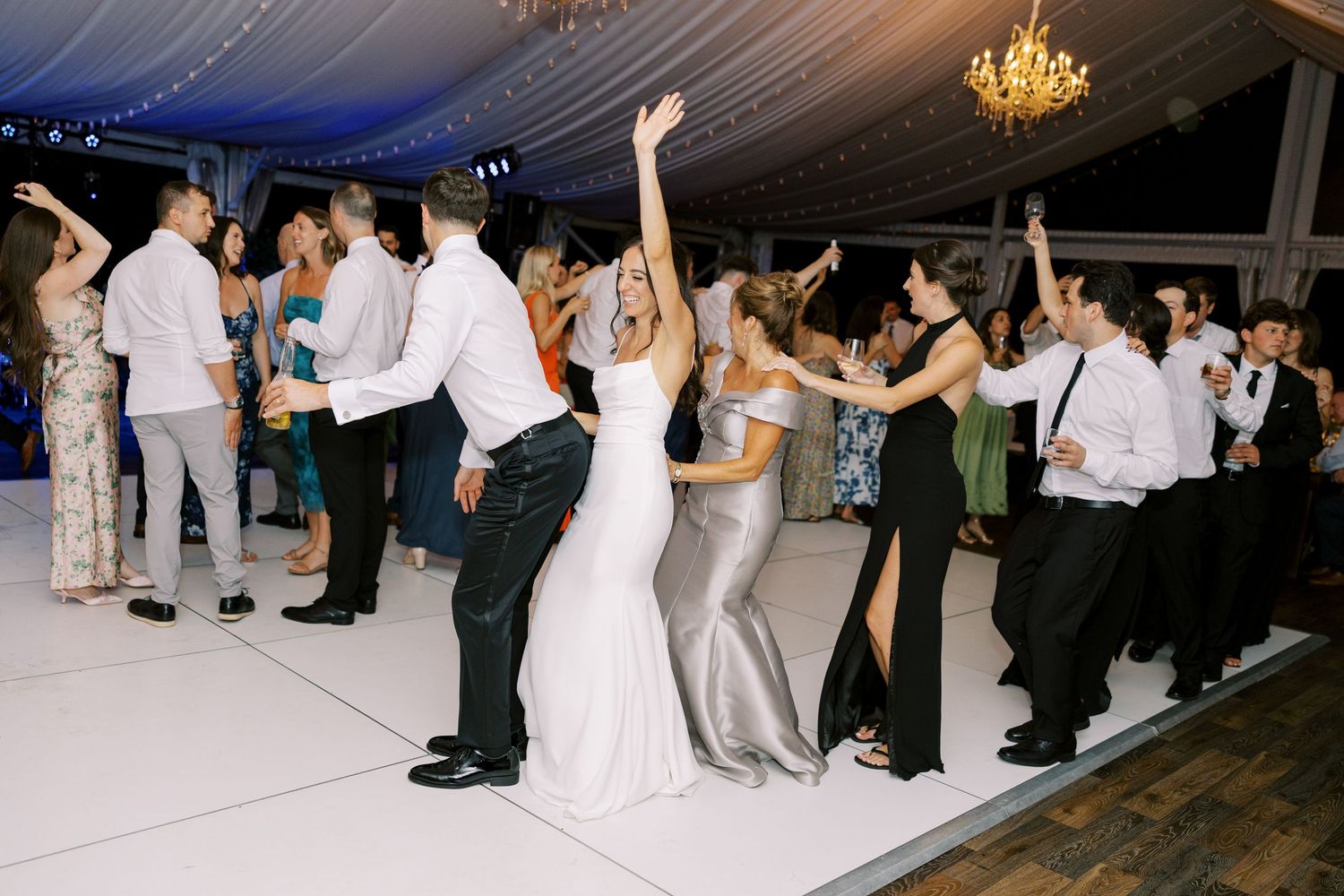Wedding guests dance under glowing chandelier during evening reception in an elegant white tented venue.