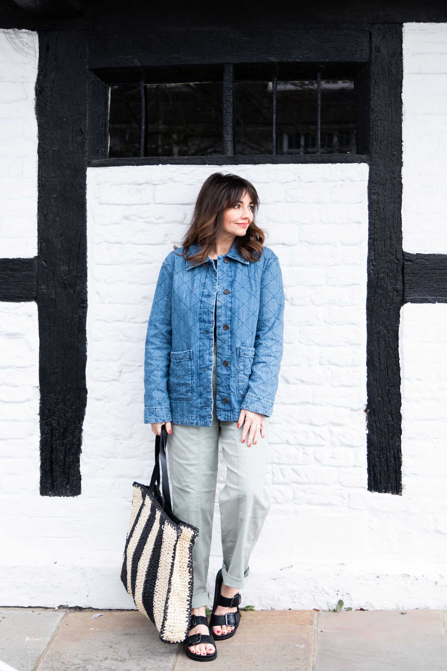 Casual spring outfit featuring quilted denim jacket, loose pants and striped straw tote bag against white painted wall.