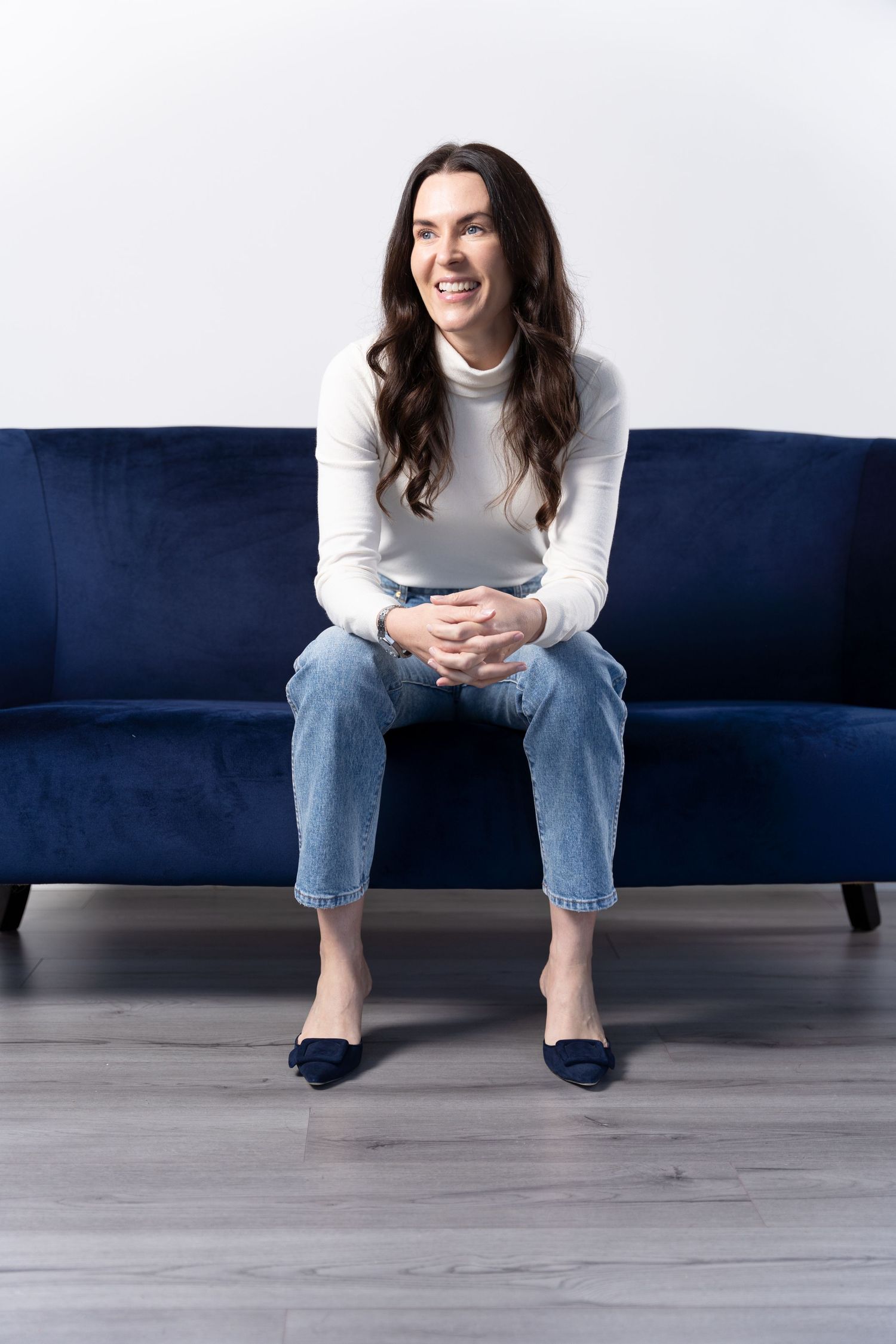Female business owner poses on a blue sofa wearing a white top during a branding photoshoot
