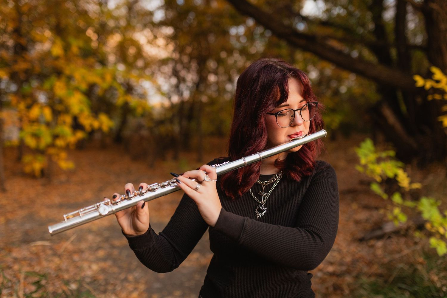 Musician plays a silver flute outdoors in a wooded area with autumn foliage during golden hour.