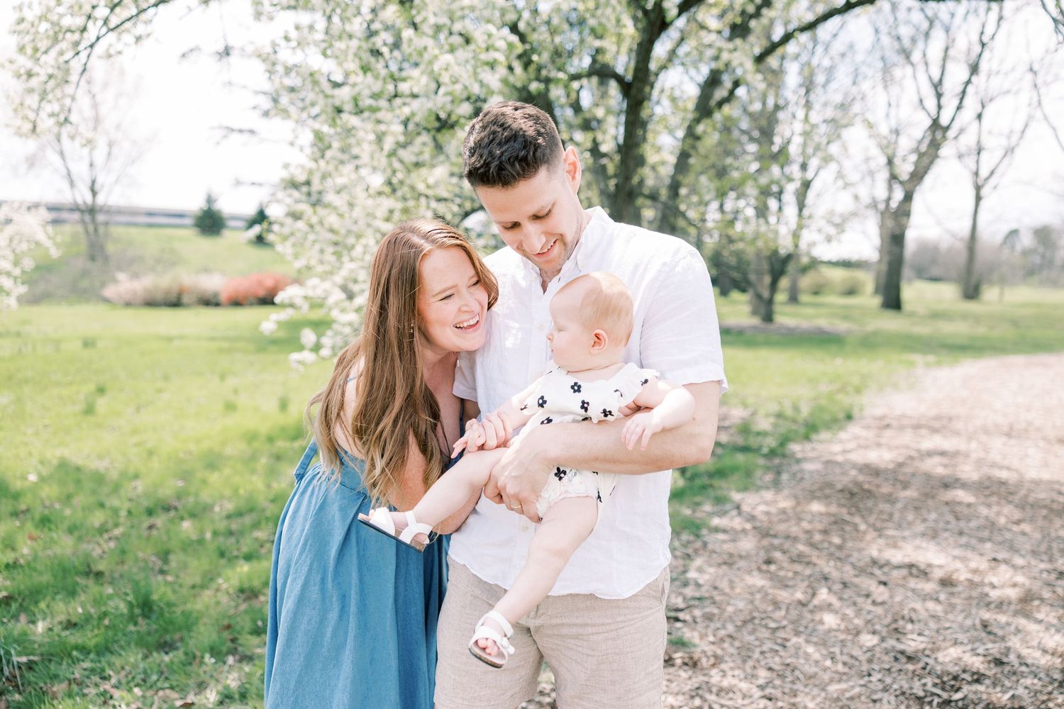 A couple stands with their baby on a spring day with blooming trees in a rural outdoor setting.