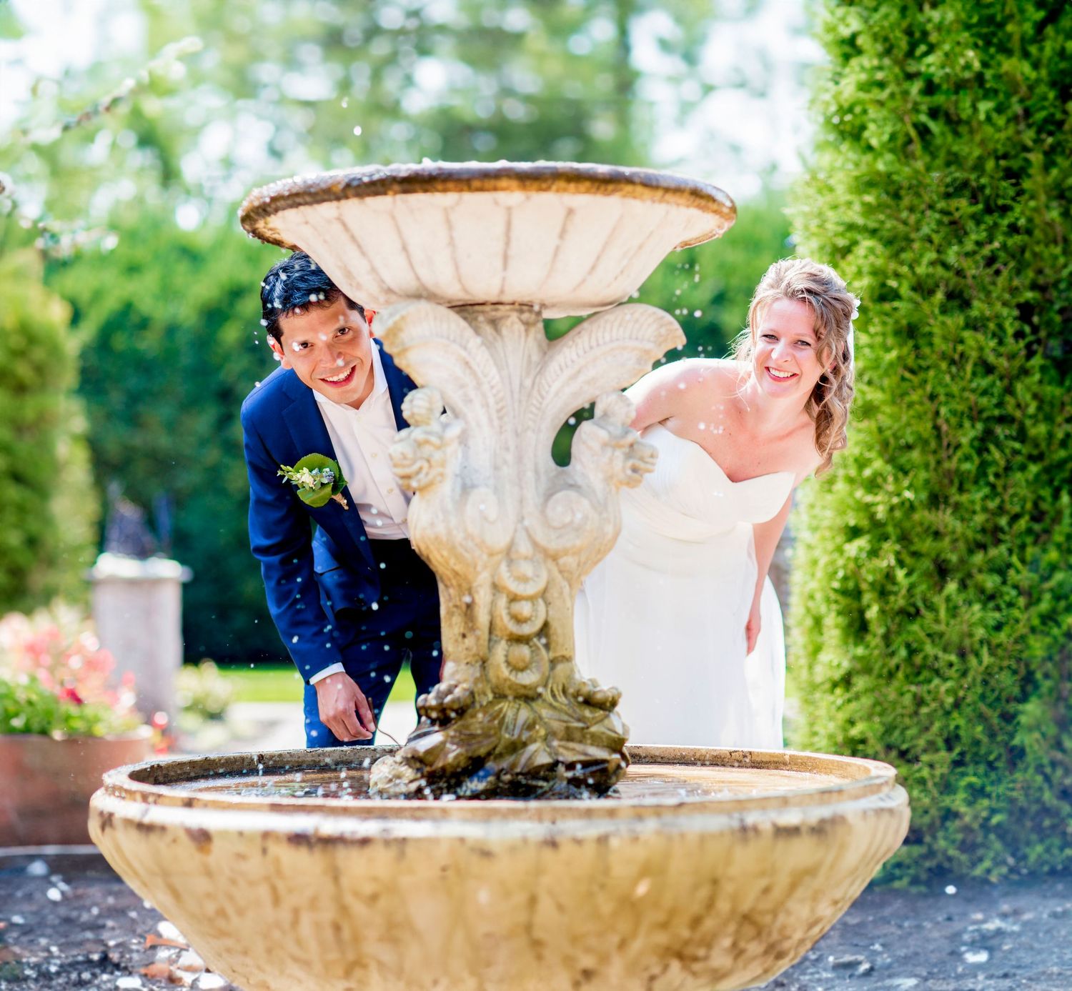 A couple in wedding attire poses playfully by an ornate stone fountain in a lush garden setting.