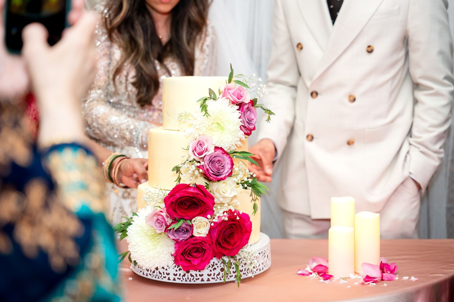 A modern three-tiered wedding cake covered in delicate lace detail and decorated with vivid red roses and pink flowers.